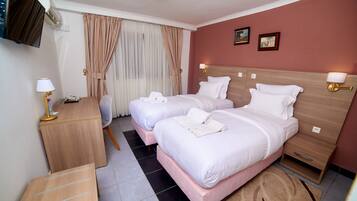 Quarto de hotel com duas camas, uma escrivaninha, uma cadeira, uma televisão e uma janela com cortinas.