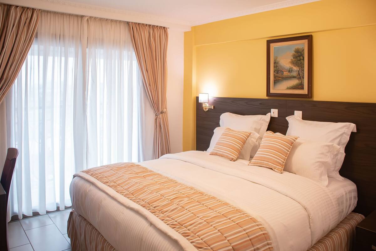 A hotel room with a large bed, a nightstand, a painting on the wall, and a window with curtains.