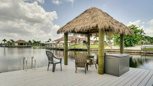 Un ponte di legno con un gazebo a tetto di paglia, una vasca idromassaggio e mobili da esterno, vicino a una superficie d'acqua tranquilla.
