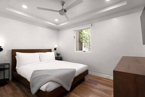 Une chambre moderne avec un lit en bois, un ventilateur de plafond, un éclairage encastré, une fenêtre donnant sur un paysage verdoyant et un petit bureau en bois.