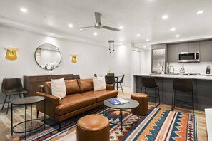A modern living room with a brown leather sofa, two black chairs, a coffee table, and a patterned rug. A kitchen area with a microwave and bar stools is visible in the background.
