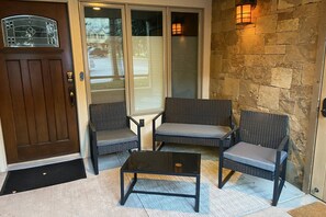 Un porche avec une porte marron, deux fauteuils en osier, une petite table et un mur en pierre agrémenté d’une lanterne.