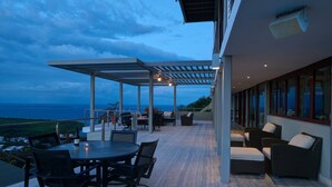 En moderne udendørs terrasse med et rundt bord og stole, et overdækket siddeområde og udsigt til havet.