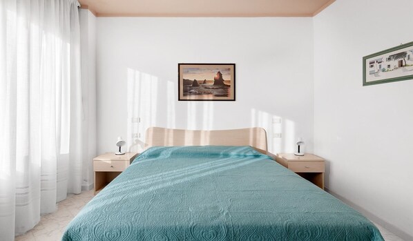 Une chambre à coucher avec un lit, deux tables de chevet, une commode, une fenêtre avec des rideaux légers et un tableau encadré au mur.
