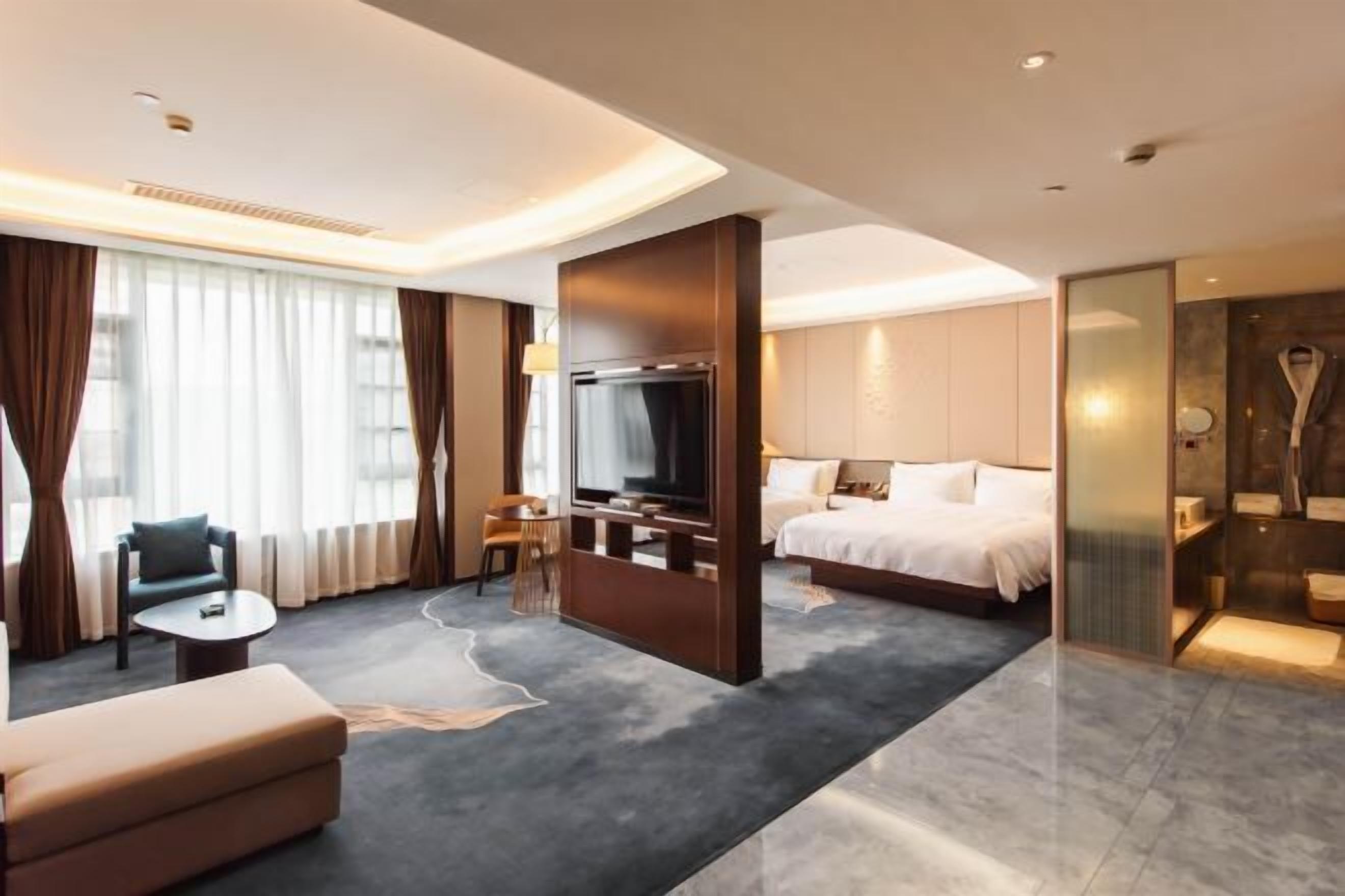 A modern hotel room with a bed, a TV, a seating area with a sofa and a small table, and a large window with curtains.