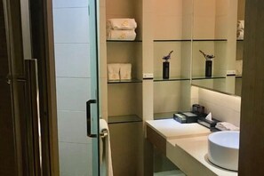 Un baño con cabina de ducha de vidrio, un lavabo blanco y repisas con toallas y artículos de aseo.