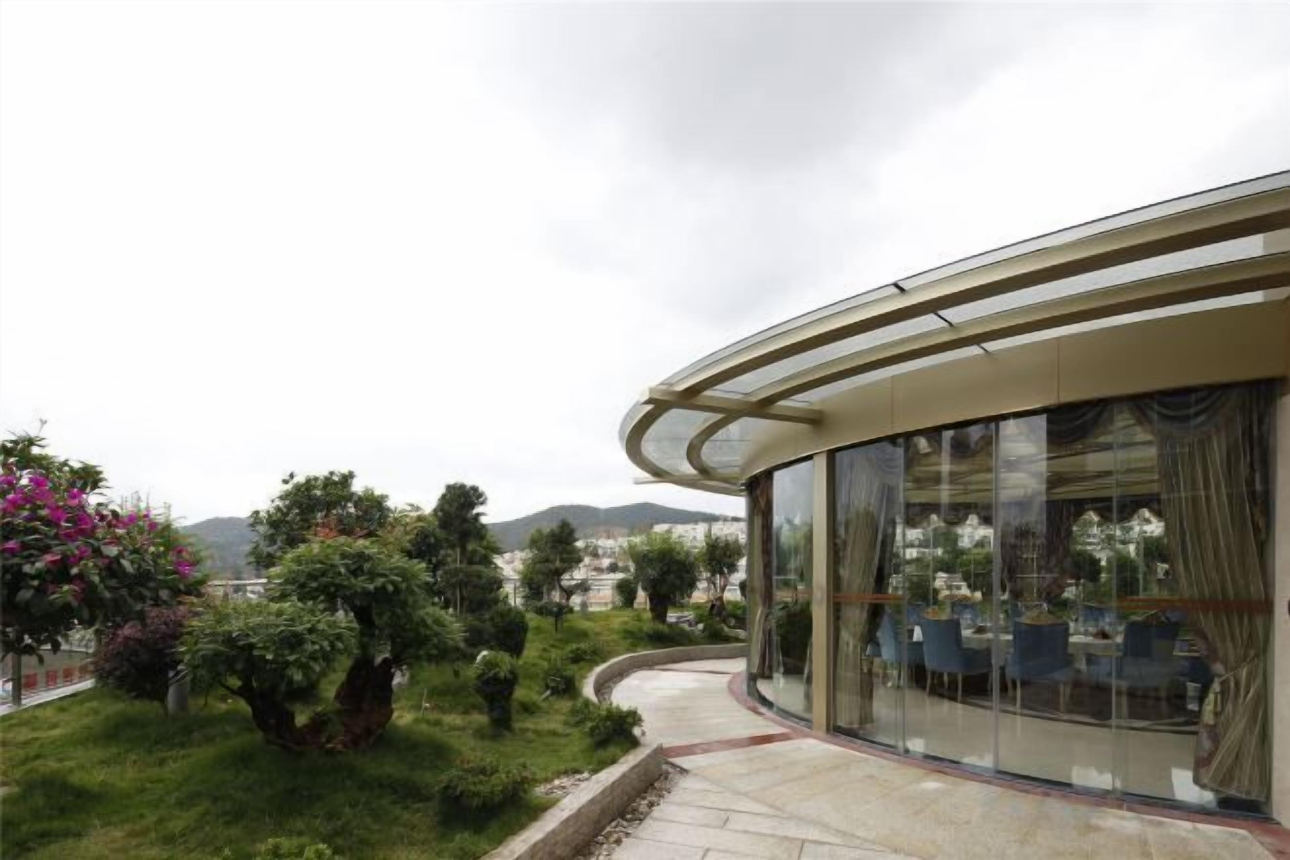 Un edificio moderno con fachada de vidrio curva, un jardín ajardinado con arbustos y flores variados, y vistas a colinas distantes.
