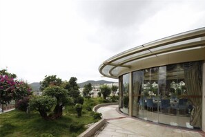 A modern building with a curved glass facade, a landscaped garden with various shrubs and flowers, and a view of distant hills. - Shanshui Hotel (Yuxi)