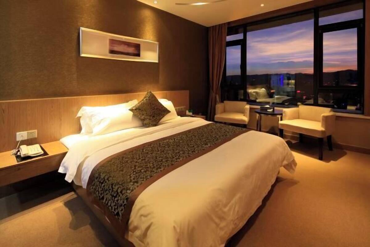 Una habitació d'hotel amb un llit gran, una taula de treball amb un telèfon, una cadira i una vista de la ciutat al capvespre.
