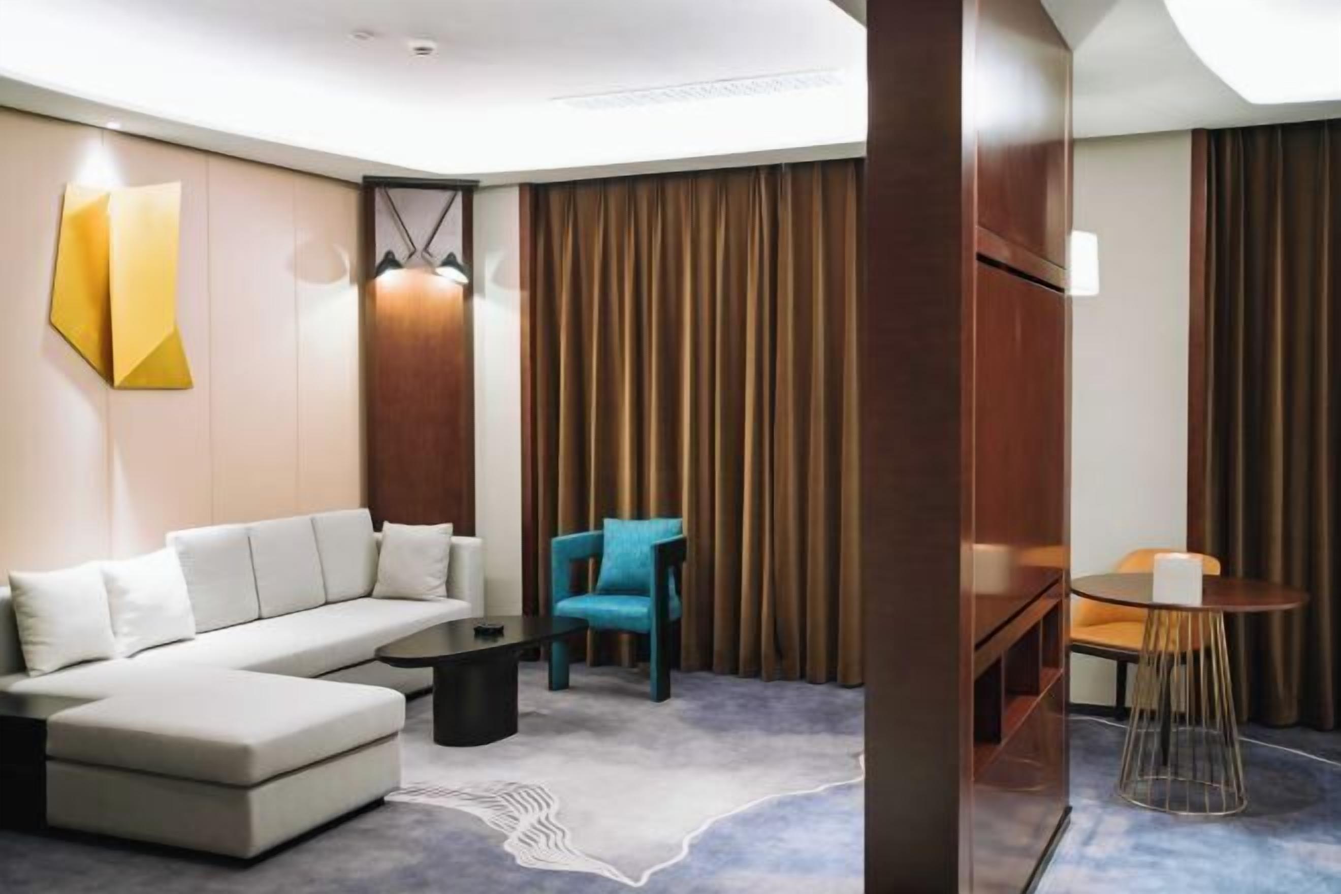 Quarto de hotel moderno com sofá modular, uma mesa redonda pequena, uma cadeira azul e uma janela grande com cortinas.