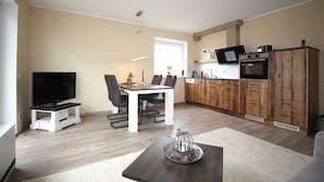 A modern living room with a flat-screen TV, dining table, and chairs. There is a kitchen area with wooden cabinets and a built-in oven. - 107-3 Holiday apartment Klabautermann - 107-3 Klabautermann (Juist)