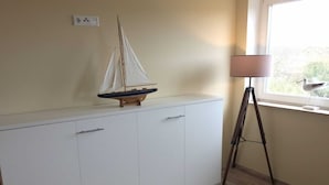 A room with a white cabinet, a small sailboat model, a lamp, and a window with a view of greenery. - 107-3 Holiday apartment Klabautermann - 107-3 Klabautermann (Juist)