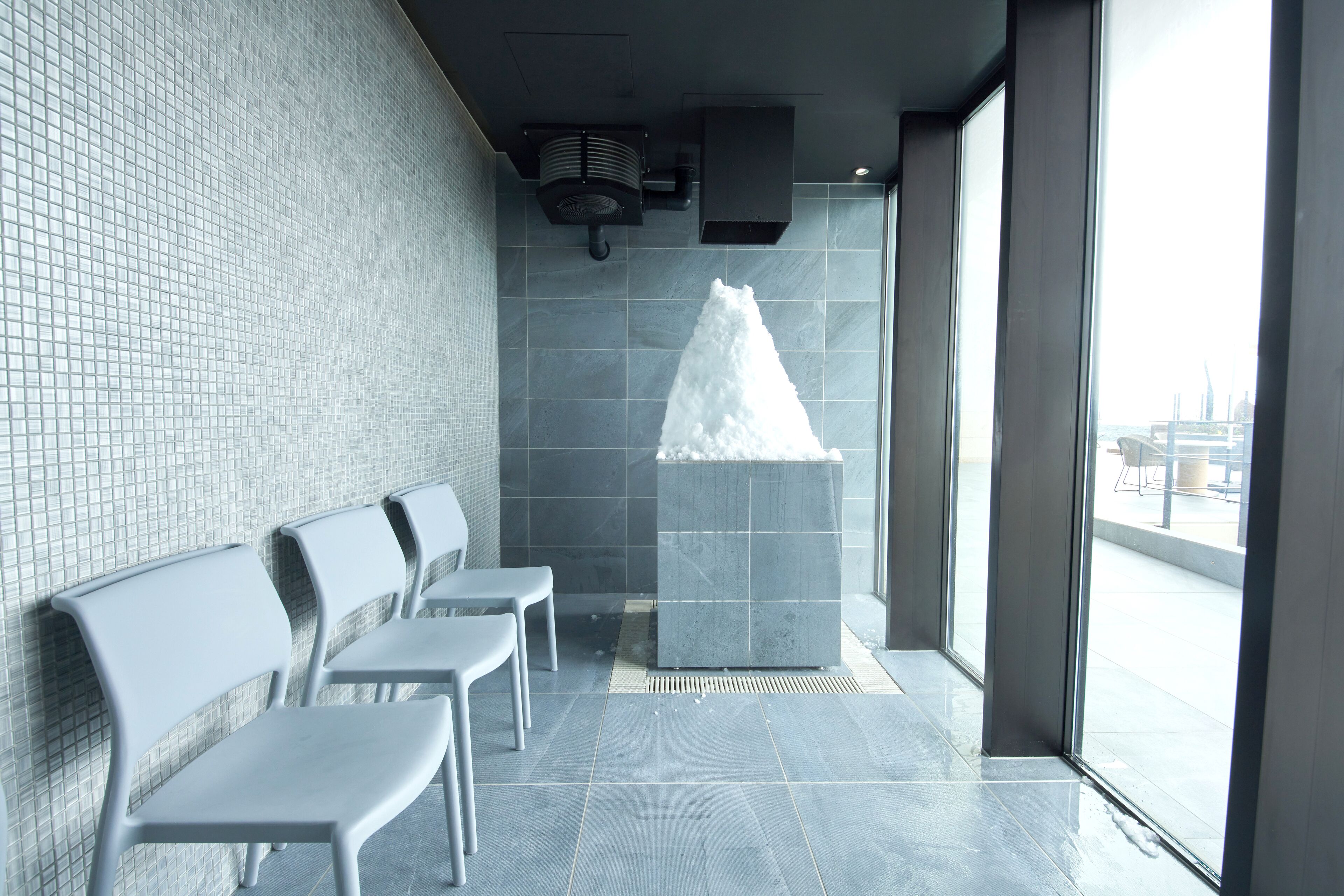 A waiting area with three grey chairs, a tiled wall, a large window, and a water feature.