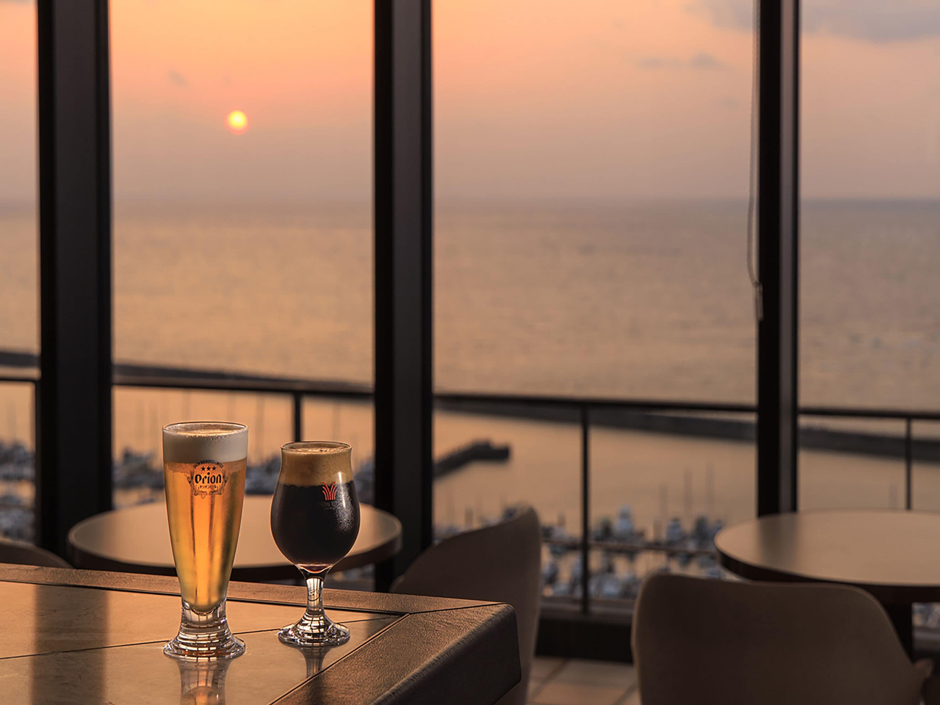 Two glasses of beer on a table with a view of the sunset over the ocean.