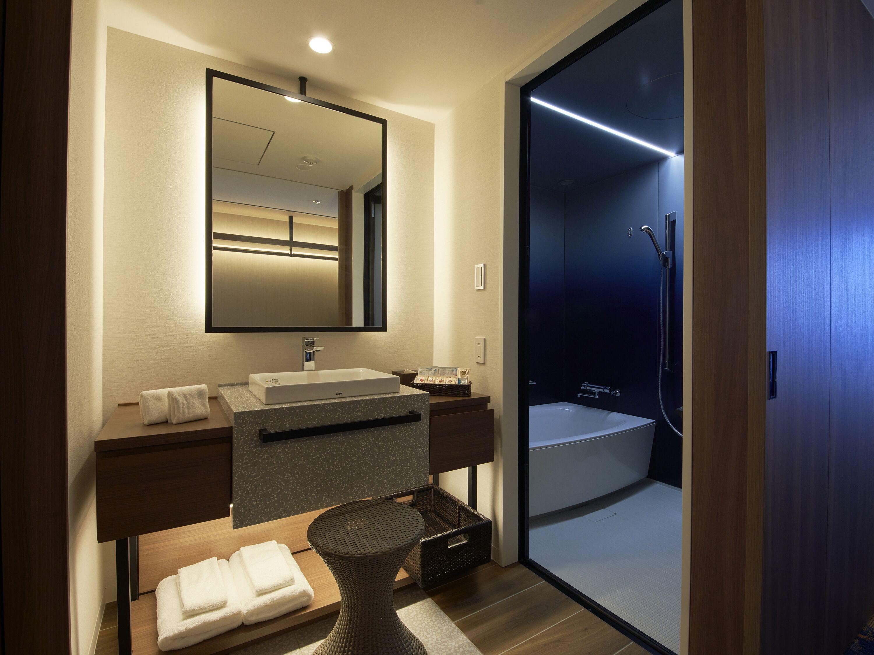 A modern bathroom with a large mirror, a rectangular sink, a shower with a glass door, and neatly folded towels on a wooden bench.