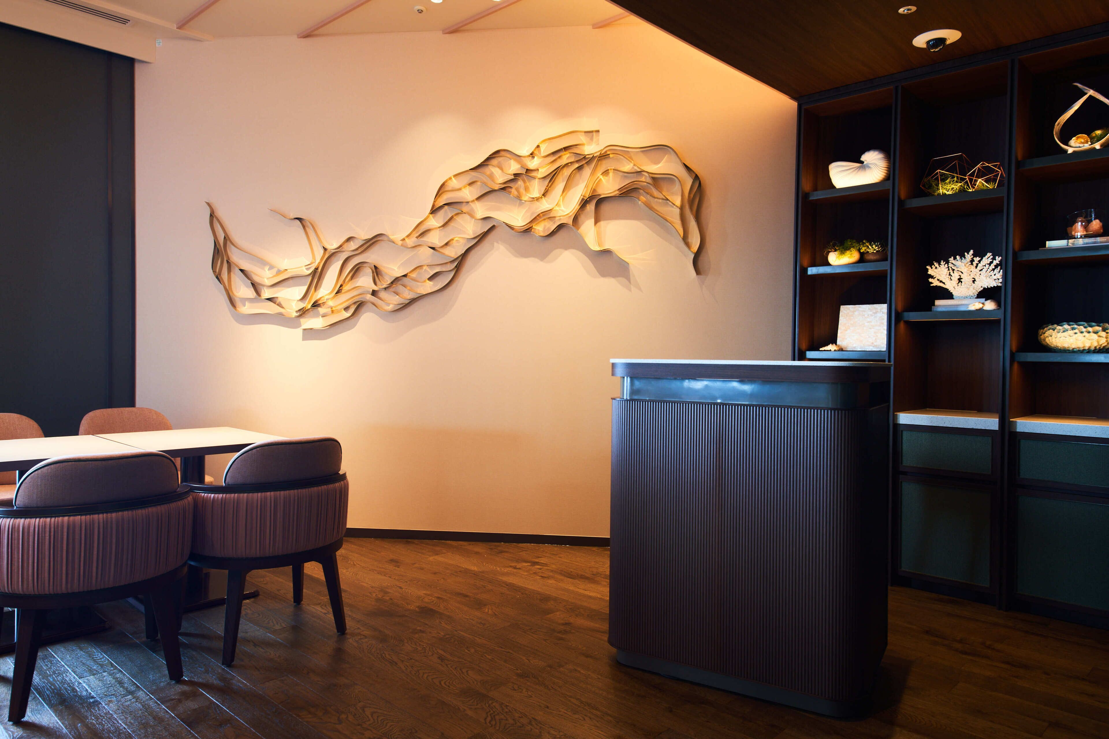 A modern interior with a wooden floor, a reception desk, a seating area with chairs, and a wall-mounted abstract art piece.