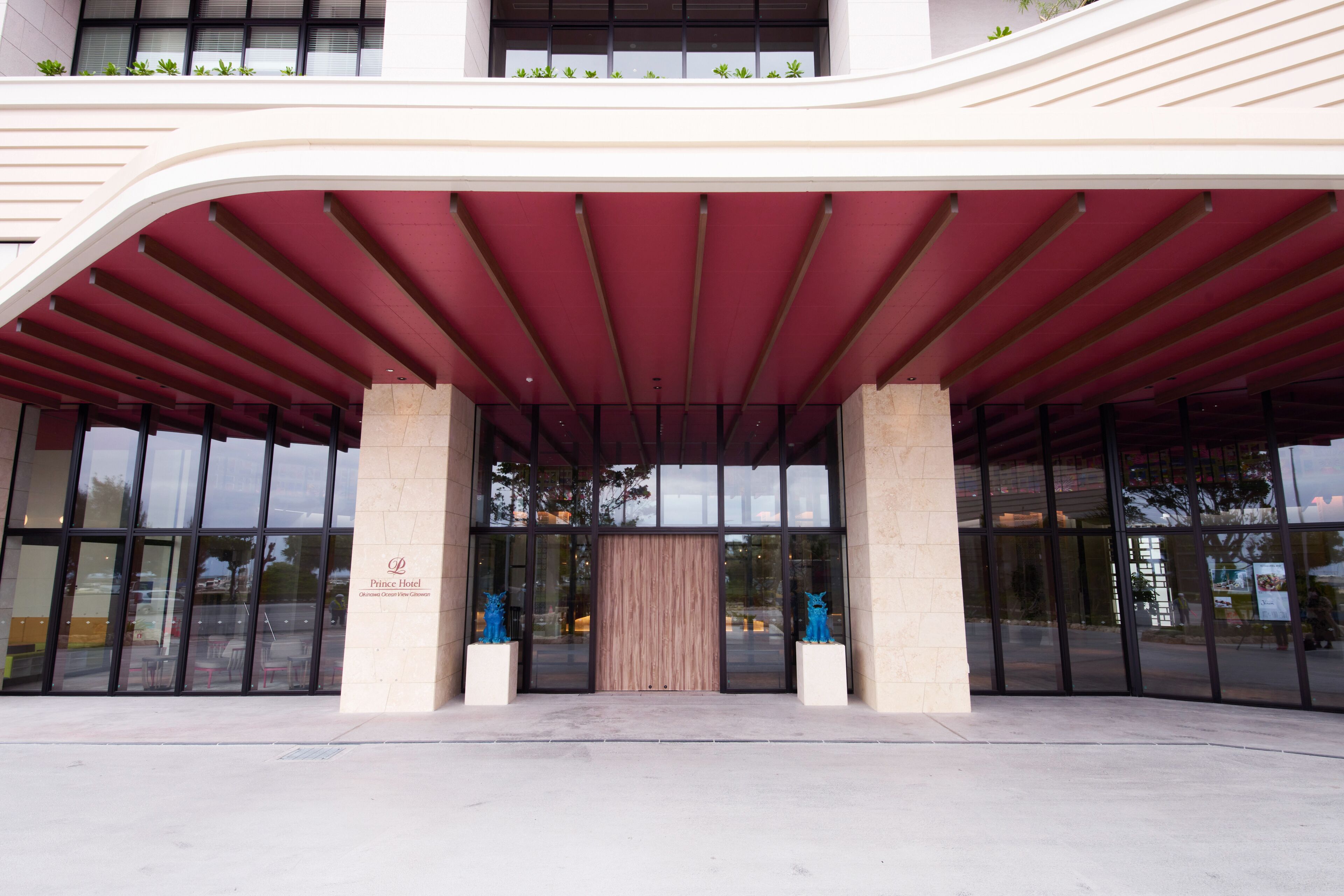 A modern hotel entrance with a curved red roof, large glass doors, and two blue sculptures on either side.