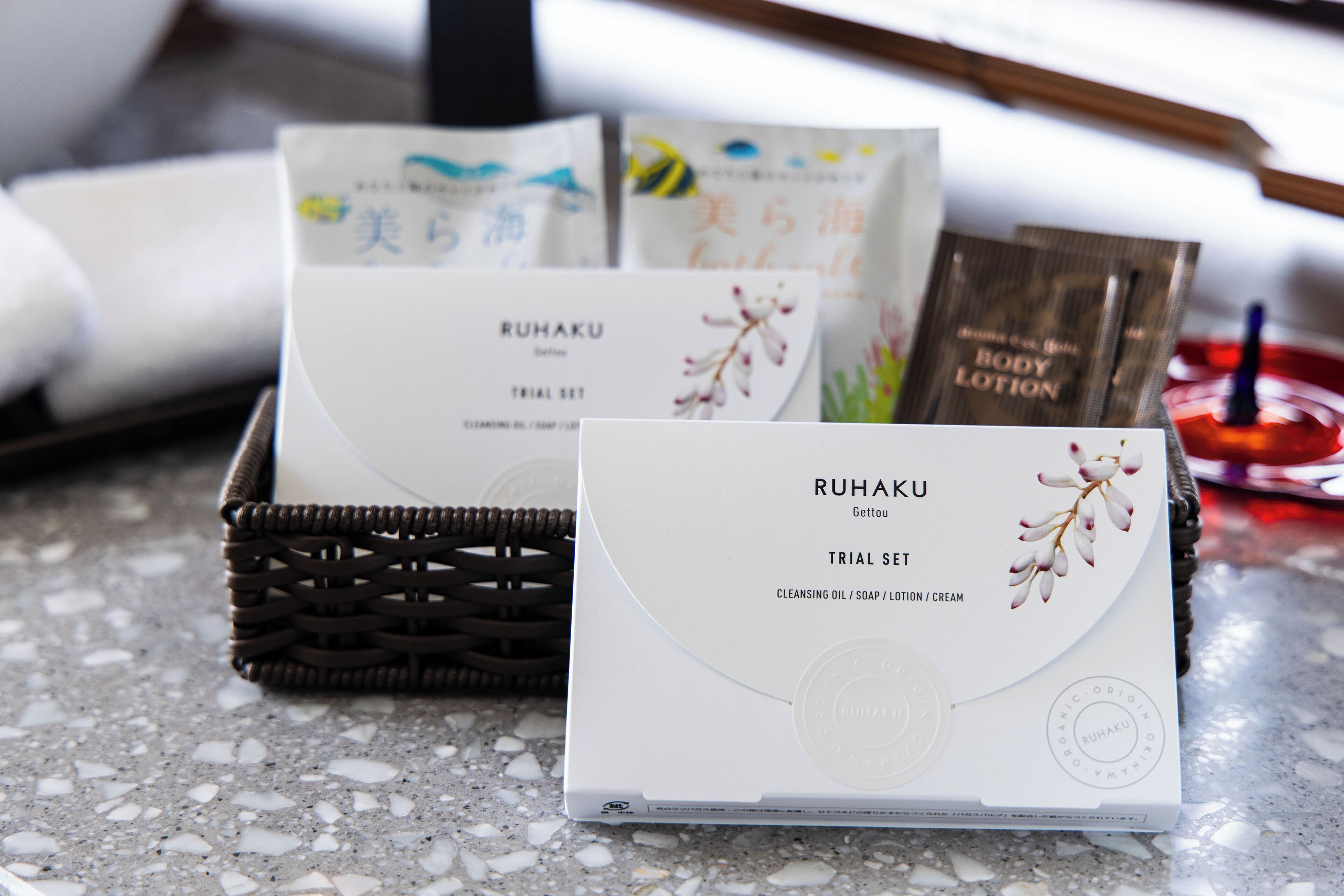 A basket containing a white box labeled 'RUHAKU' with 'TRIAL SET' and 'CLEANSING OIL / SOAP / LOTION / CREAM' text, and a card with similar branding.