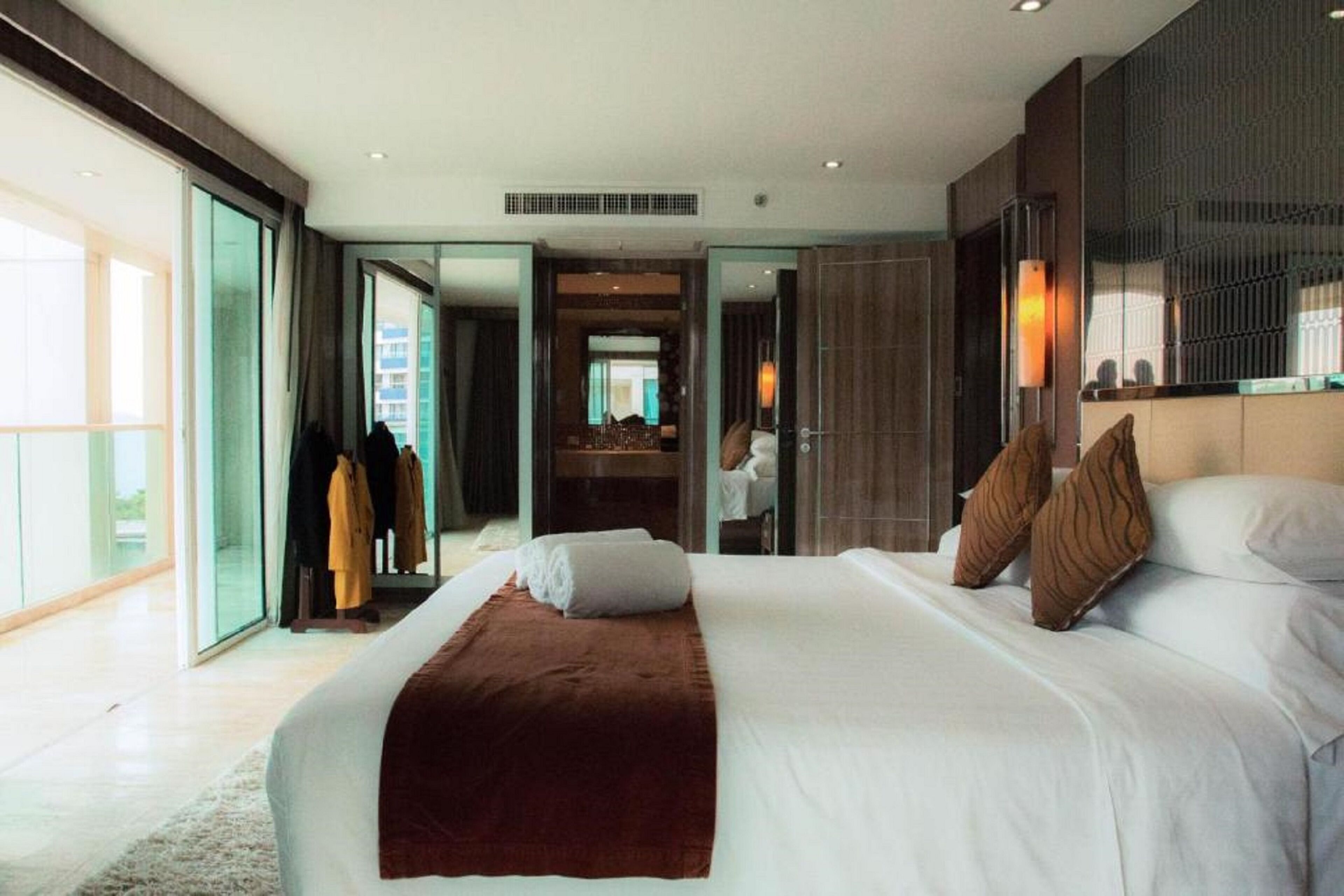 Une chambre d’hôtel moderne dotée d’un grand lit, d’un dressing et offrant une vue sur la ville grâce à de grandes fenêtres du sol au plafond.