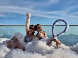 Two people in a bubble bath with a clear view of the sea and sky.