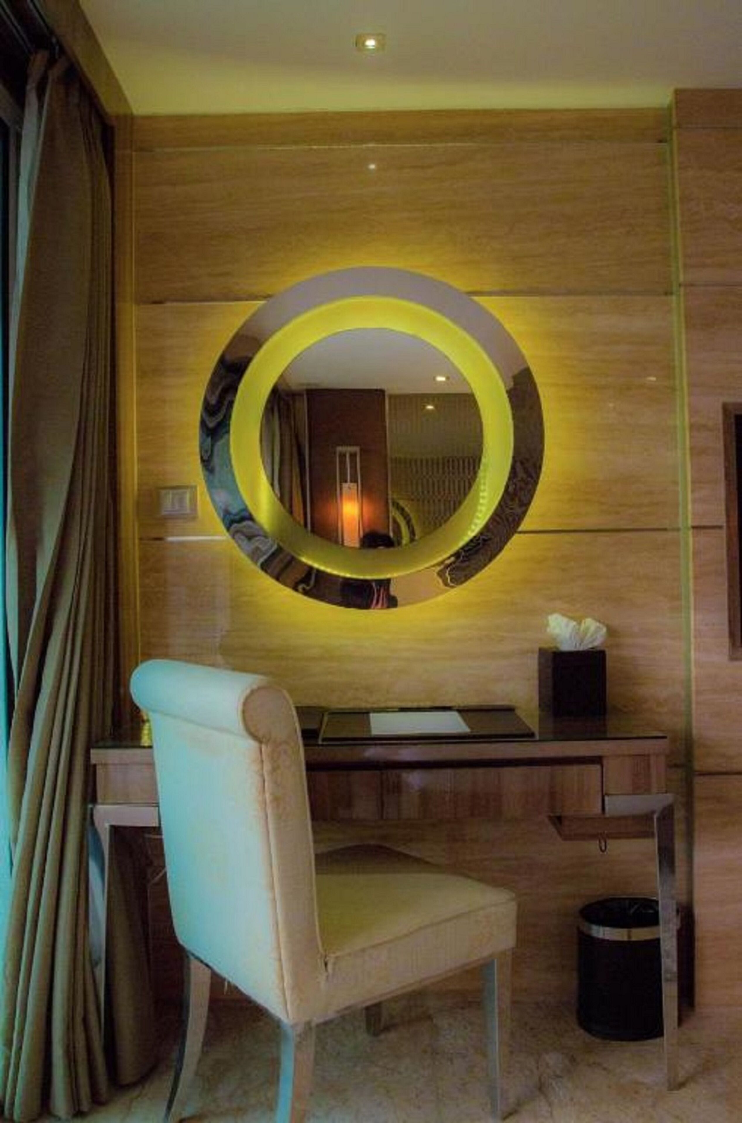 A hotel room with a wooden wall, a round mirror with a yellow frame, a desk, a chair, and a trash bin.