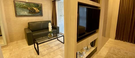 Una sala de estar moderna con un sofá de cuero negro, una mesa de centro de vidrio con un jarrón de flores blancas, un televisor de pantalla plana montado en un panel de madera y un cuadro de un paisaje fluvial en la pared.