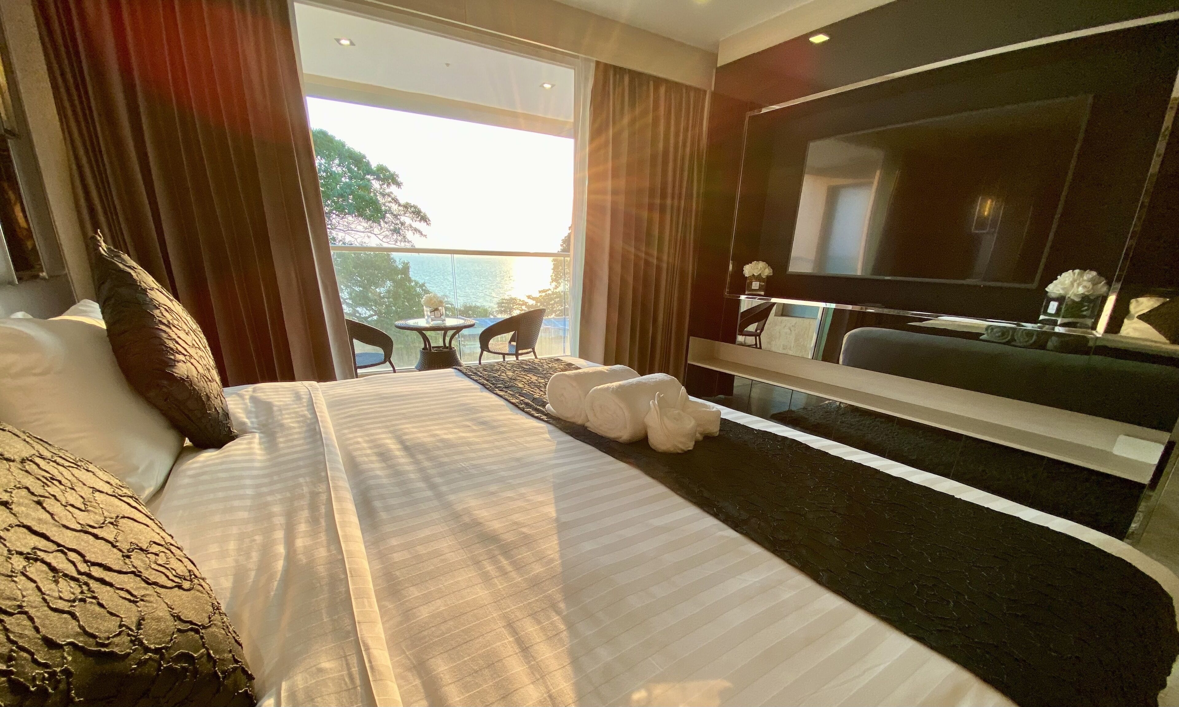 Una habitación de hotel moderna con una cama grande, un televisor de pantalla plana, un balcón con vista al mar y una pequeña mesa con sillas.
