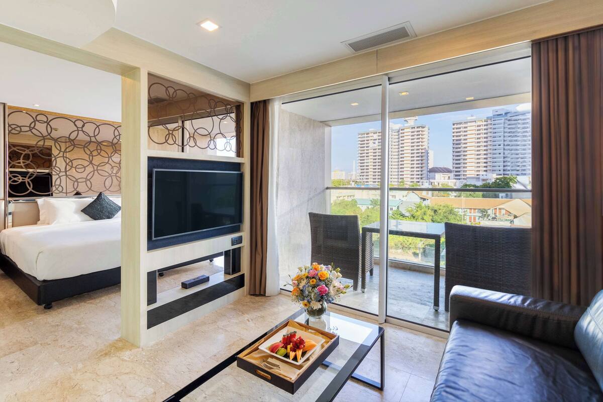 Een moderne hotelkamer met een bed, een flatscreen televisie, een eettafel met een fruitplateau, een bank en een balkon met uitzicht op hoogbouw.