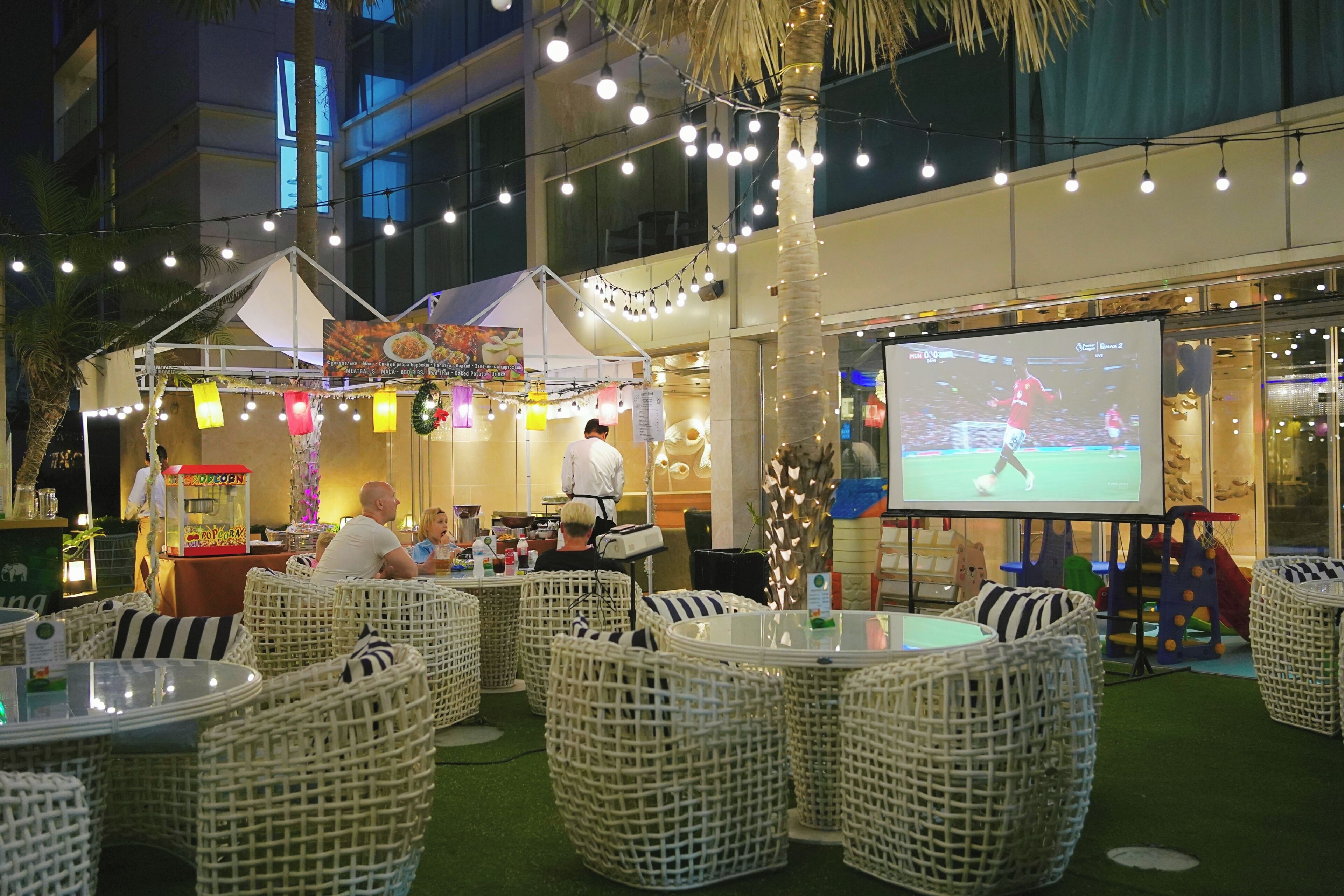 Outdoor seating with wicker chairs and tables, a food kiosk, and a large screen displaying a sports event.