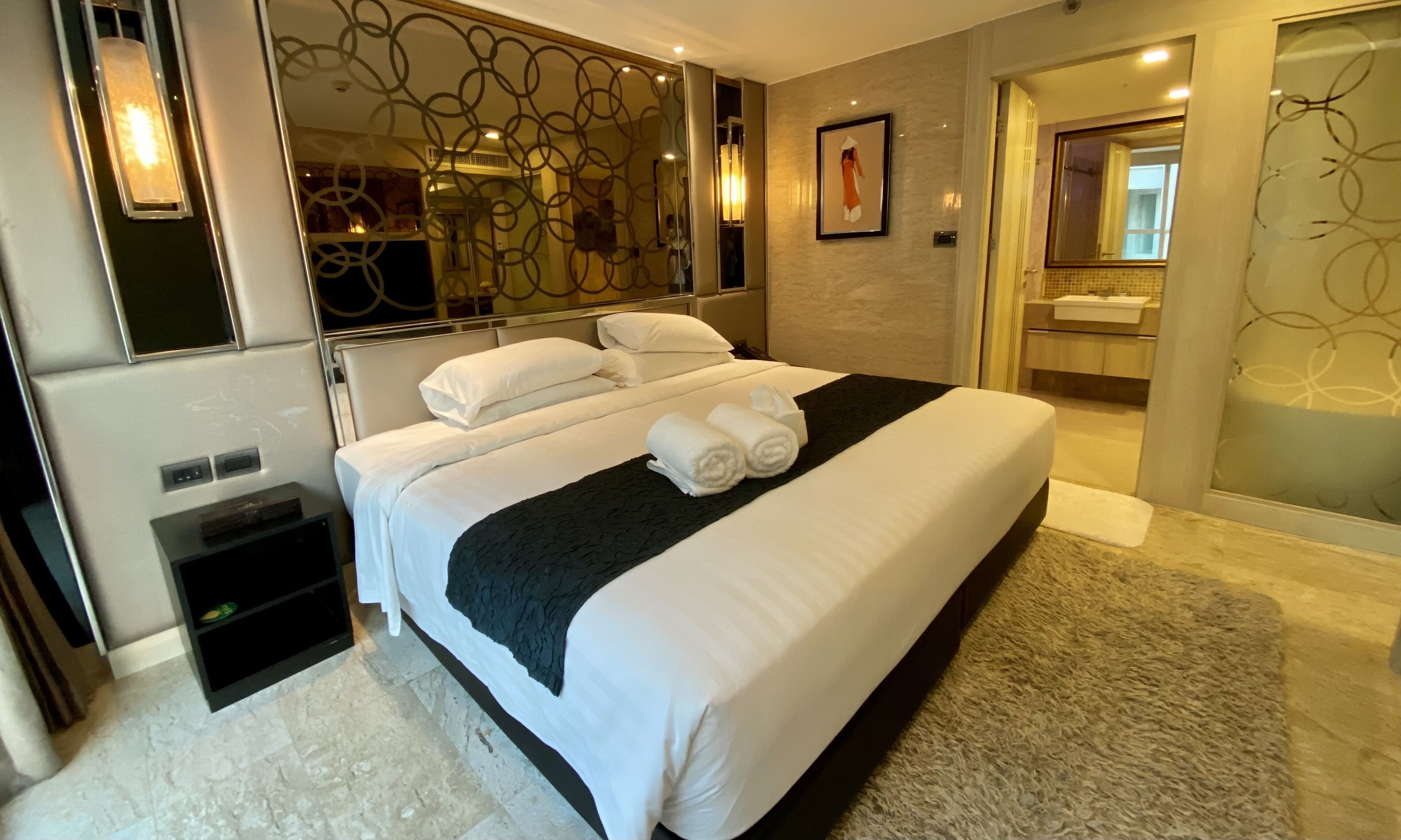 A hotel room with a large bed, a black throw blanket, and two rolled towels. There is a large mirror on one wall and a bathroom visible through an open door.