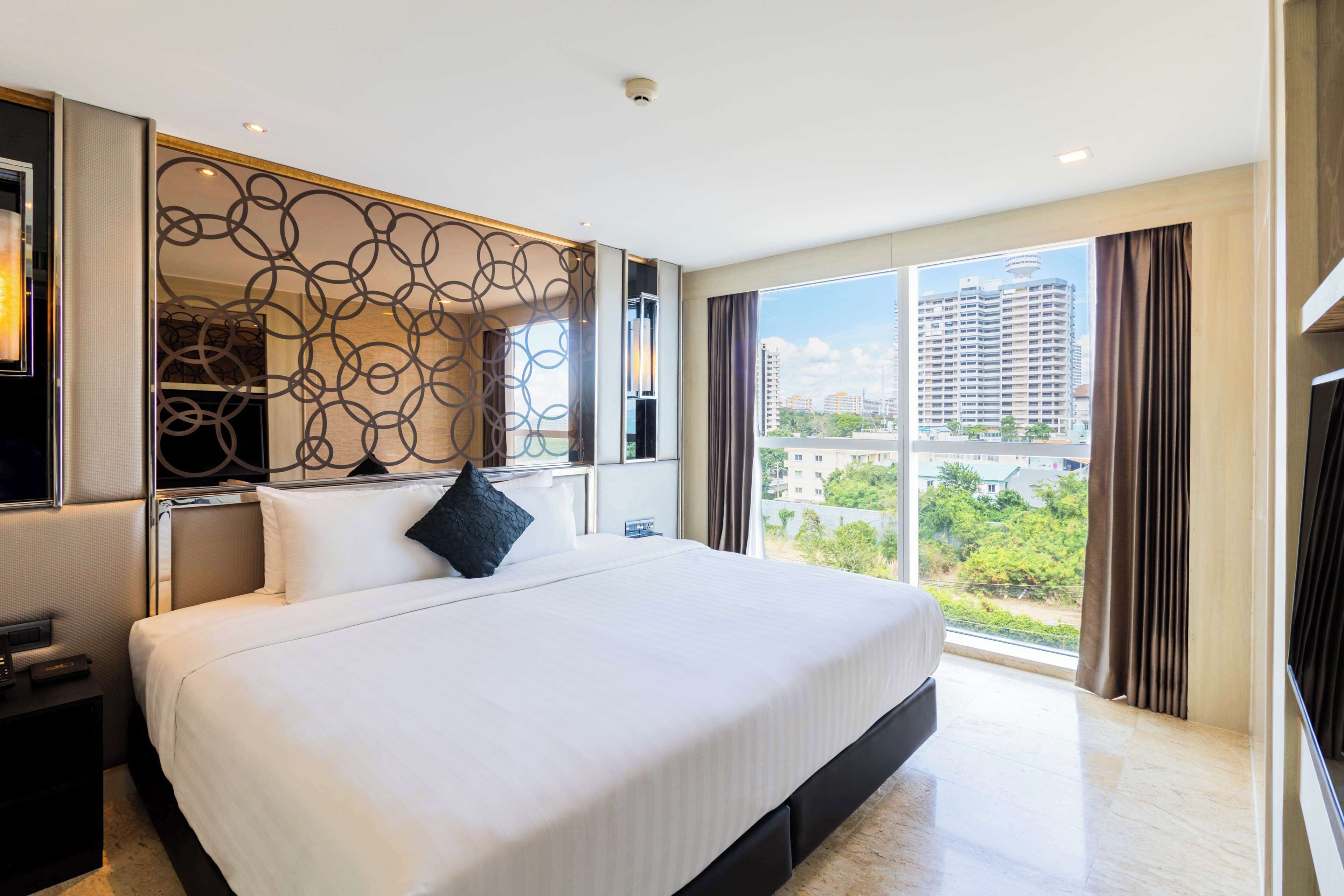 Een moderne hotelkamer met een groot bed, een decoratief wandpaneel, een nachtkastje en uitzicht op de stad door grote ramen.