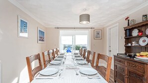 Un coin repas avec une longue table dressée pour six personnes, des chaises en bois, un lustre et une vue dégagée sur l’extérieur grâce à une grande fenêtre.