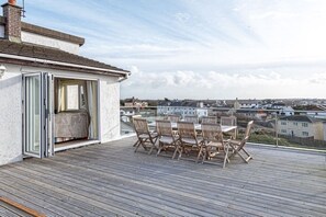 A wooden deck with a table and chairs, a sliding glass door, and a view of houses in the distance. - Trearddur Bay Home with a View, Beach & Snowdon Mountain Range and Hot Tub (Trearddur Bay)