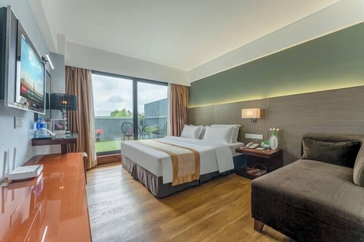 A hotel room with a bed, a desk with a TV, a chair, and a view of the outdoors.