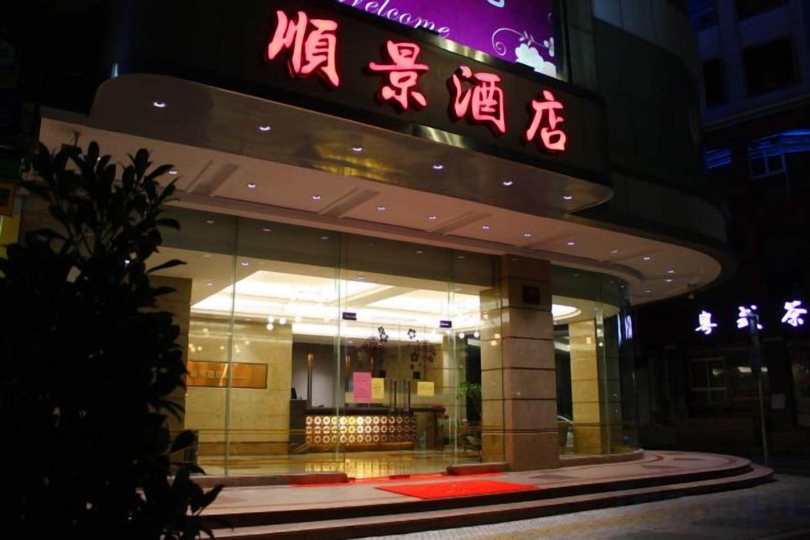 A hotel entrance with a glass facade, illuminated signage, and a red carpet leading to the door.