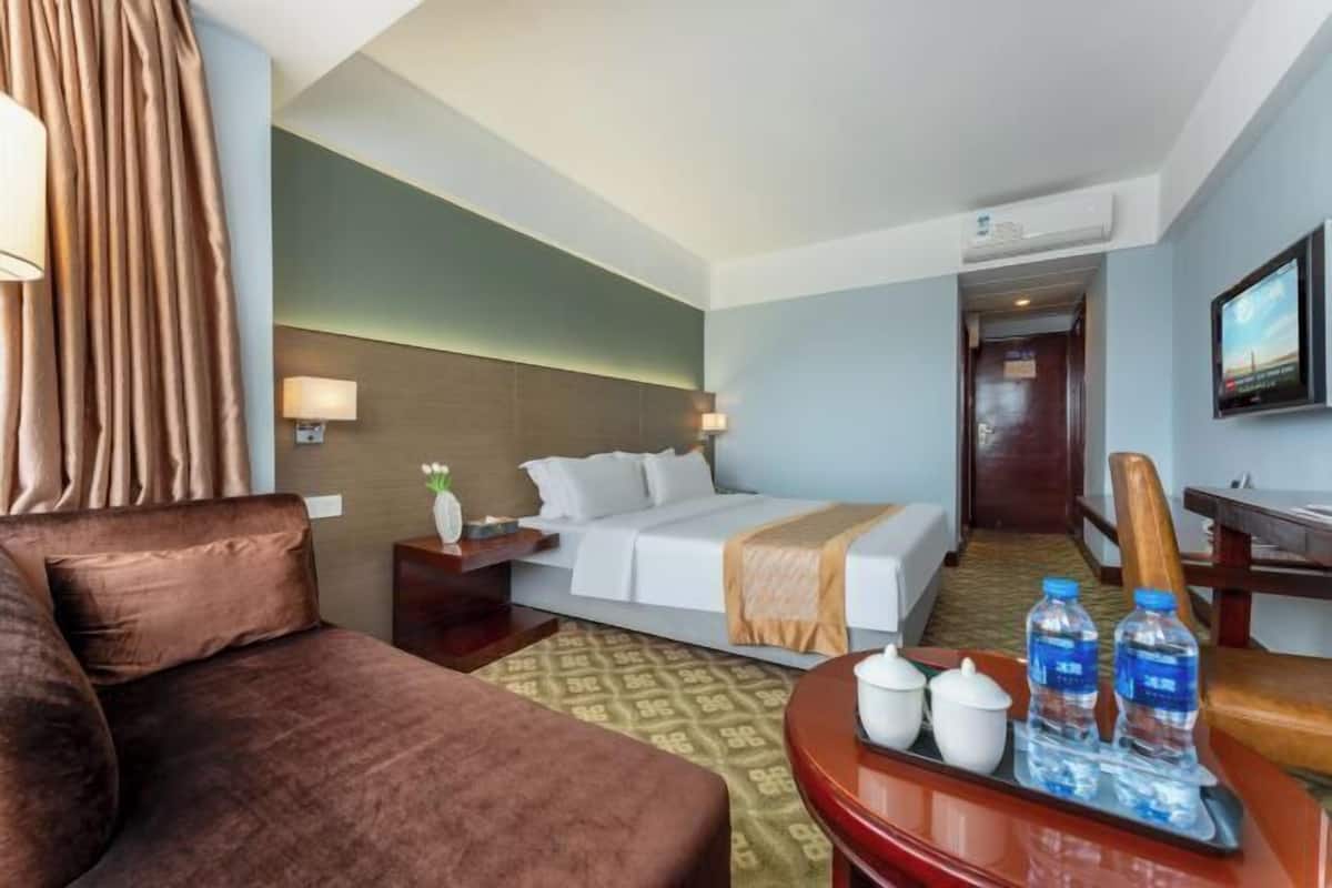 A hotel room with a bed, a sofa, a desk, and a TV. There are water bottles and a tray with cups on the desk.