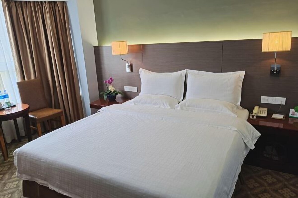 A hotel room with a large bed, two bedside lamps, a nightstand with a phone, a small table with a chair, and a window with curtains.