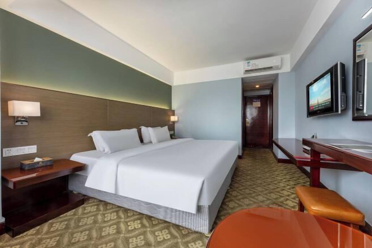A hotel room with a bed, bedside table, lamp, TV, and a desk with a chair.