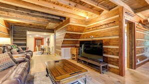 A log cabin interior with a wooden ceiling, a leather sofa, a TV, and a dining area.