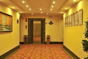 A hallway with a carpeted floor, an elevator, a painting on the left, and a wall with informational signs.