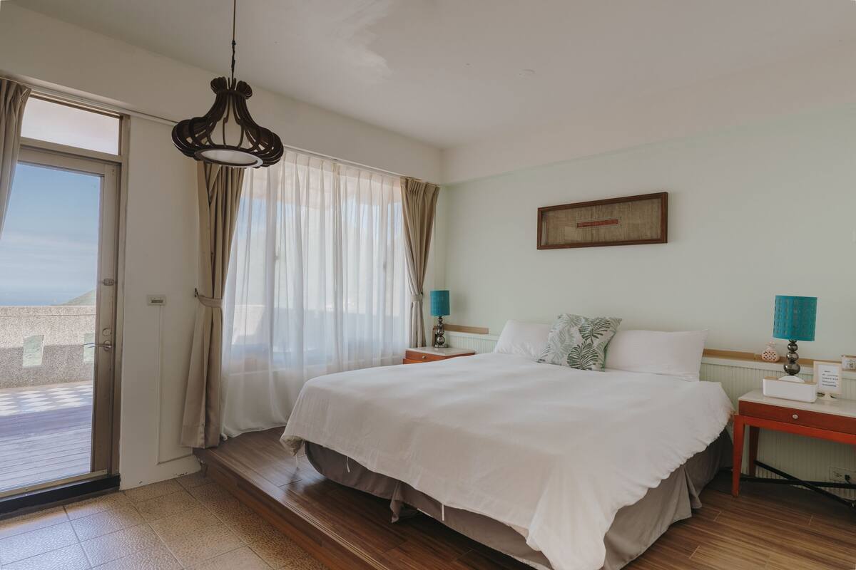 A bedroom with a large bed, a wooden floor, a sliding glass door to a balcony, and a ceiling light fixture.
