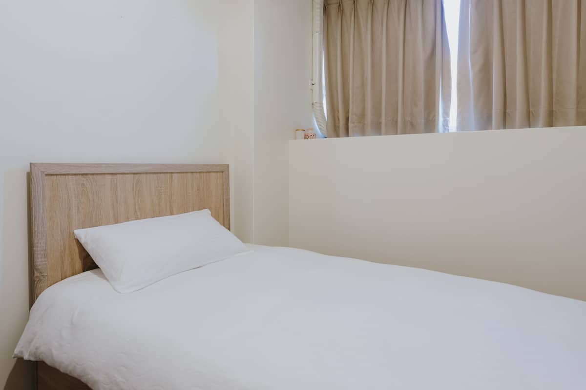 A single bed with a wooden headboard and white bedding in a room with a plain wall and a window with beige curtains.