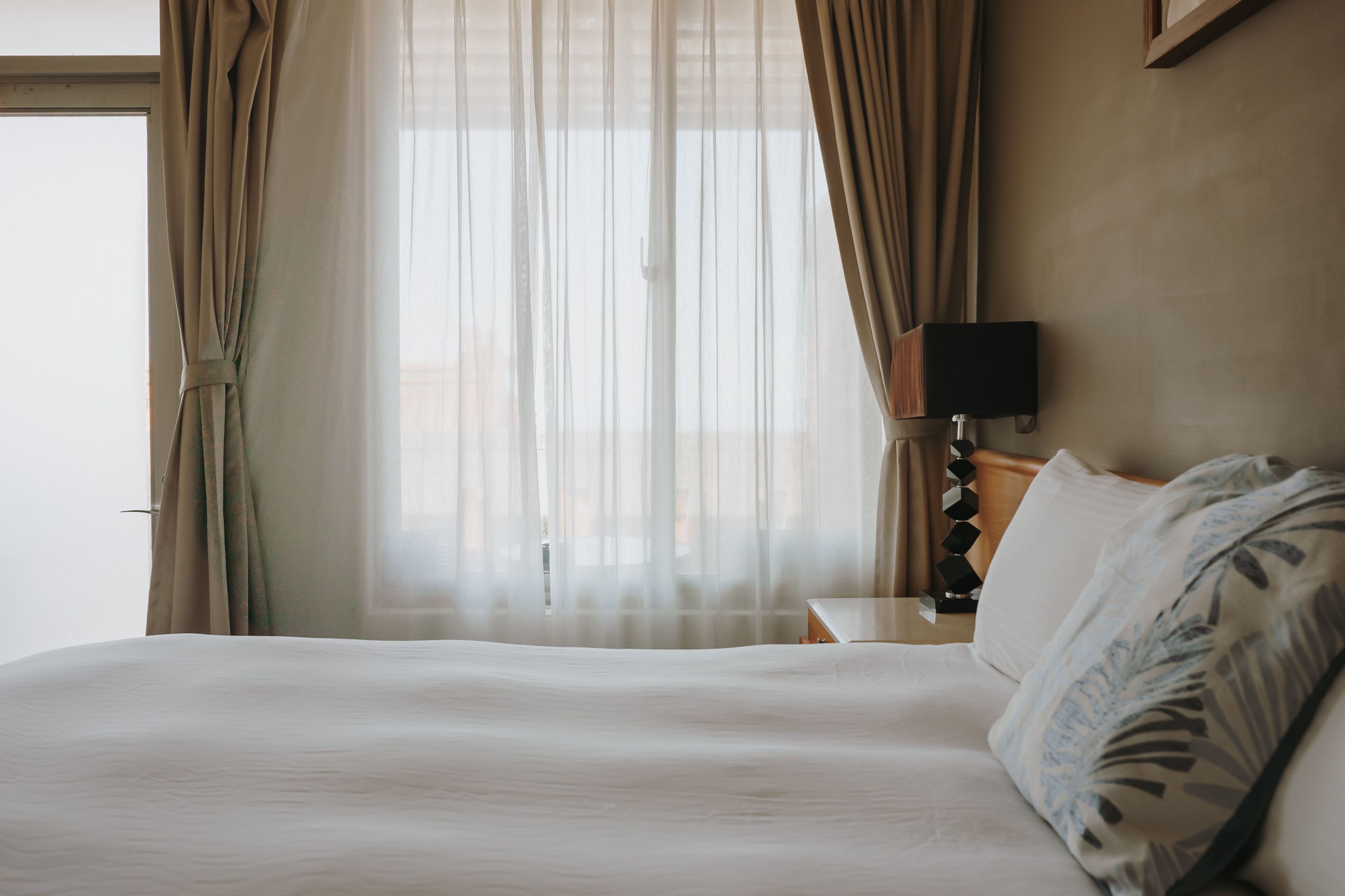Ein Hotelzimmer mit einem großen Bett, einem Nachttisch mit Lampe und durchsichtigen Vorhängen.