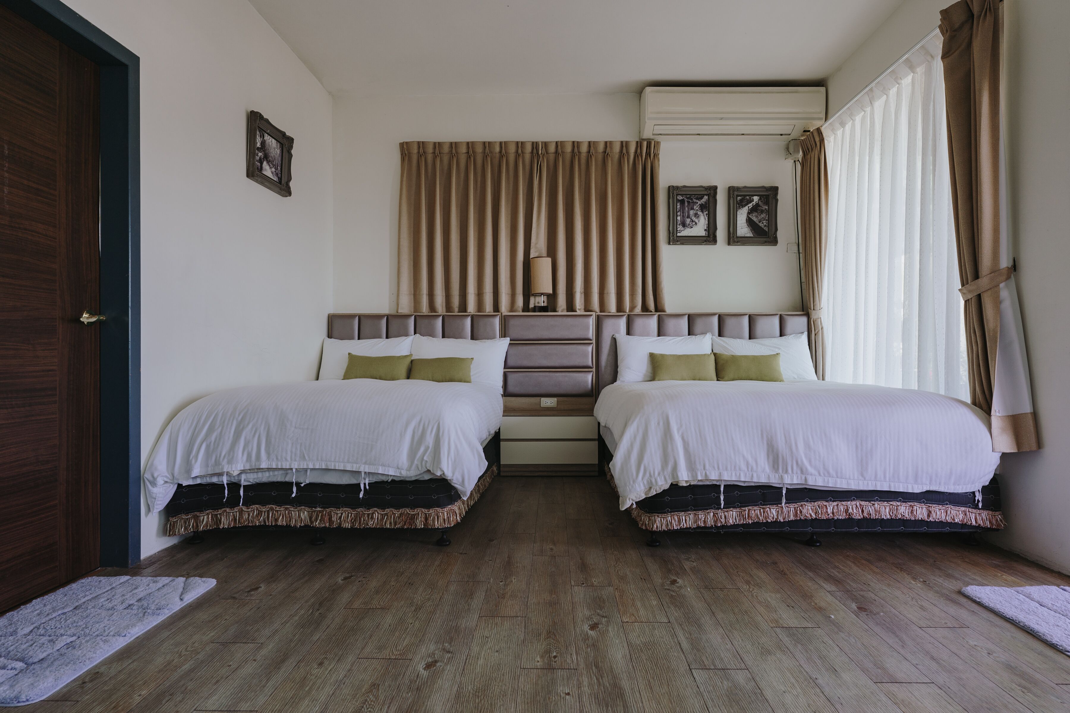Una habitación con dos camas, piso de madera, una puerta y una ventana con cortinas.