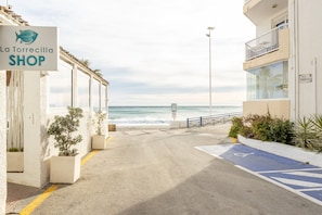 A coastal street with a shop named 'La Torrecilla SHOP' on the left, a clear view of the sea, and a blue handicapped parking space on the right.