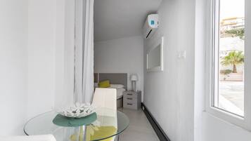 A modern hotel room with a glass table, a bed with a green pillow, a bedside lamp, and a wall-mounted air conditioner.