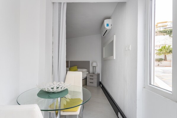 A modern hotel room with a glass table, a bed with a green pillow, a bedside lamp, and a wall-mounted air conditioner.