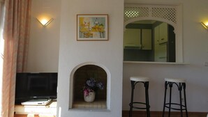 A living room with a fireplace, a framed picture, a TV, and two bar stools. - Apartment 'Les Fonts 83' with Shared Pool, Private Terrace and Balcony (L'Estartit)