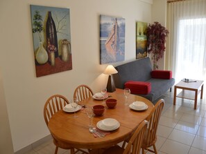 Un comedor con una mesa de madera dispuesta para cuatro personas, rodeada de sillas. Hay pinturas en las paredes, un sofá con un cojín rojo y una mesita con una lámpara.
