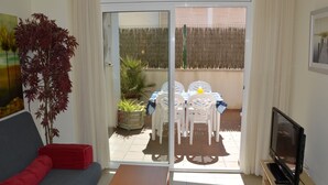 En stue med sofa, sofabord, tv på et stativ og en skydedør, der fører ud til en udendørs terrasse med bord og stole.